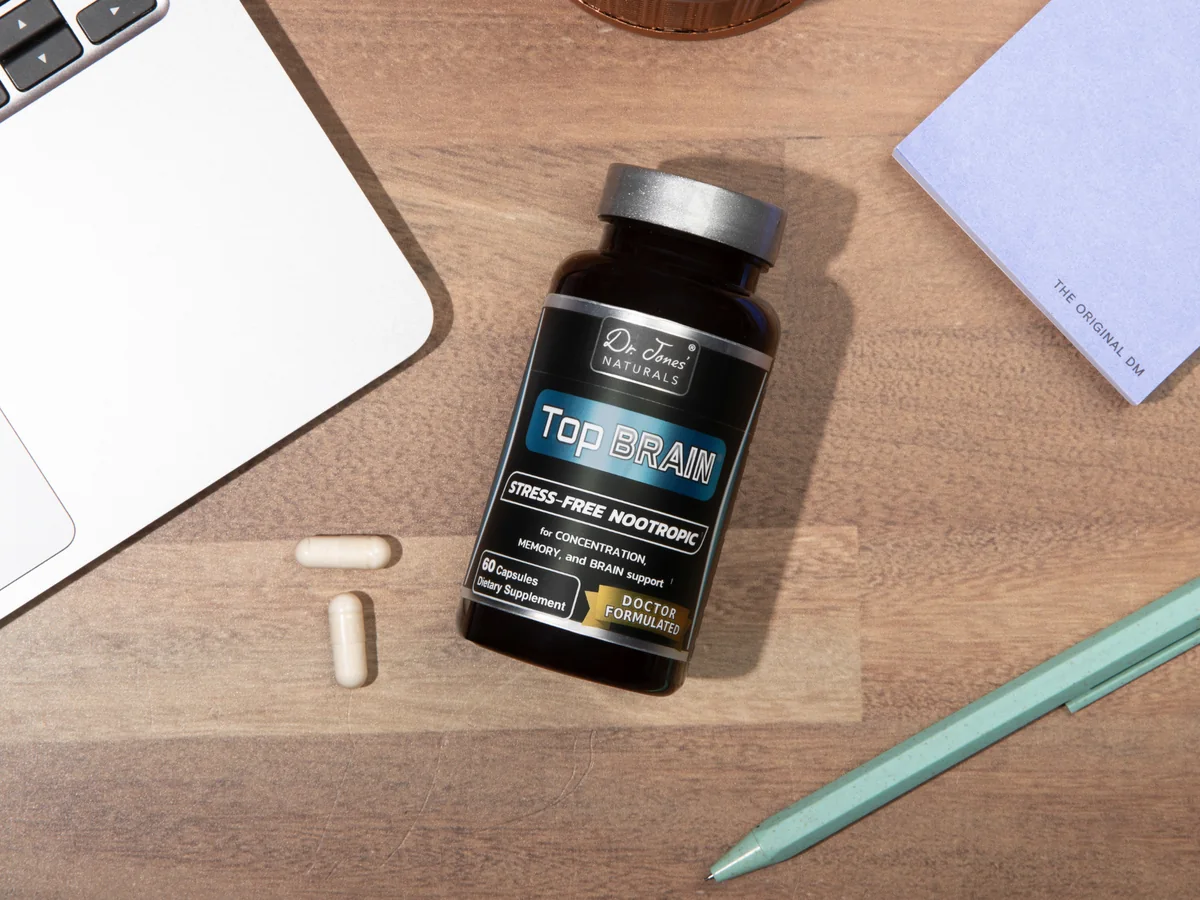 A bottle of Top Brain and two capsules on a wooden desk beside a laptop and a notepad